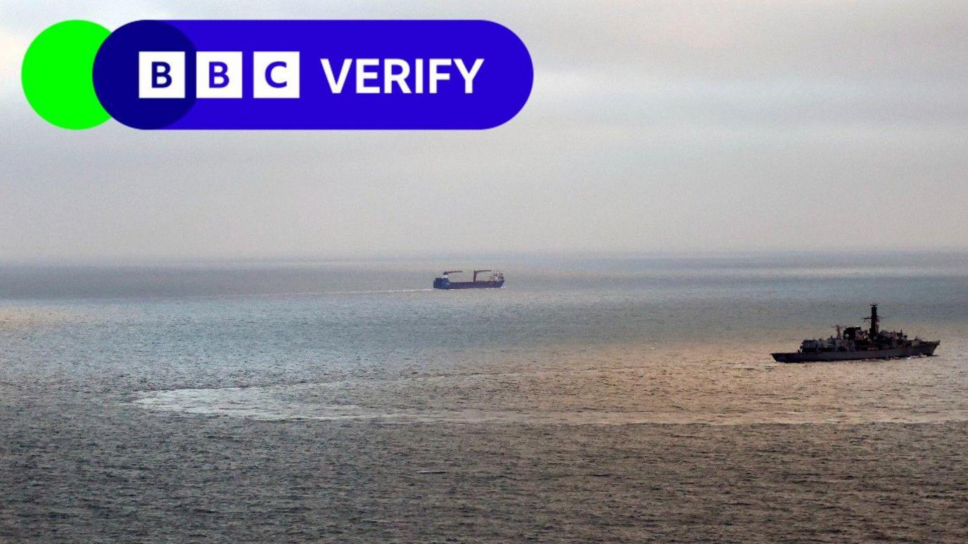 A photos of two ships. One military vessel can be seen on the right of the photo, while a commercial vessel is seen in the distance. The weather is calm and sunny. 