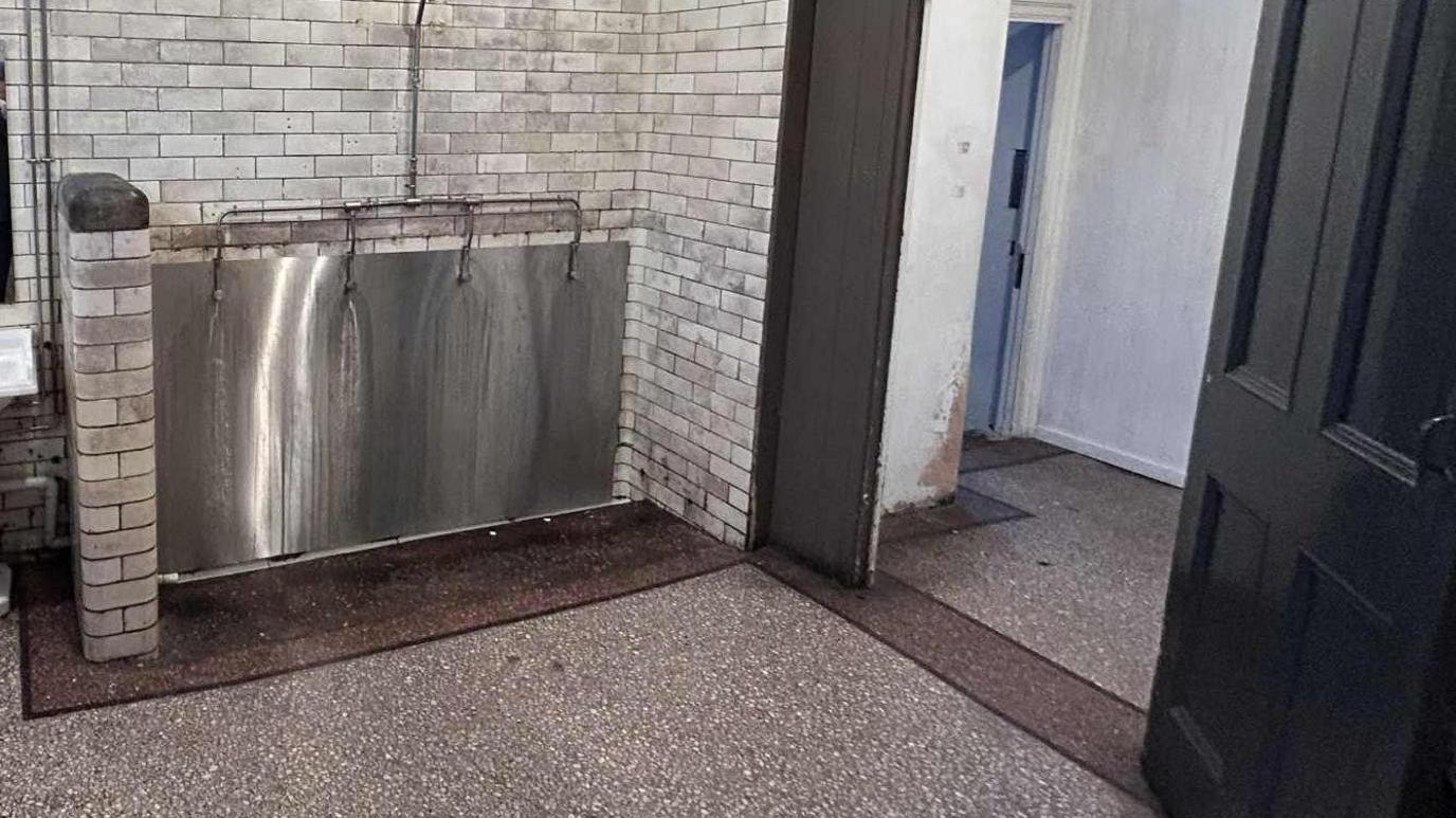 The silver urinal which is in an alcove surrounded by the grey tiles which look to be turning black in places. The floor around it looks dark. The entryway can be seen to the right, with the white painting chipped.