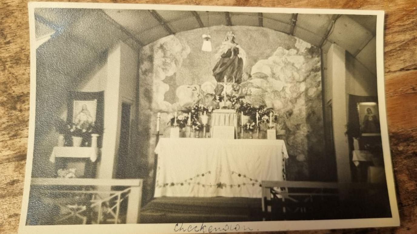 A picture of a printed black and white photo showing an alter in a World War Two-era Nissen hut, with a curved ceiling.