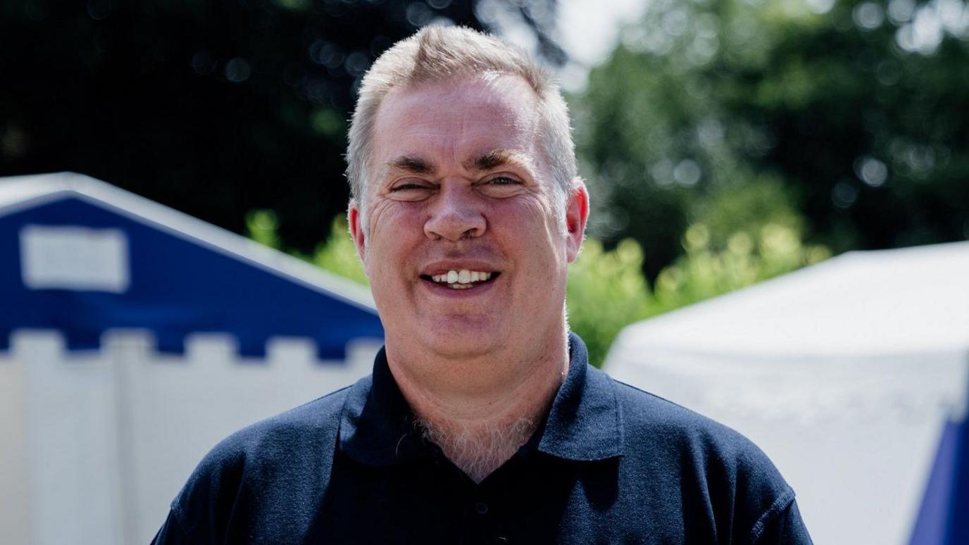 Mark Lawrie, a man with grey hair and wearing a black polo shirt, smiling at the camera with trees behind him.