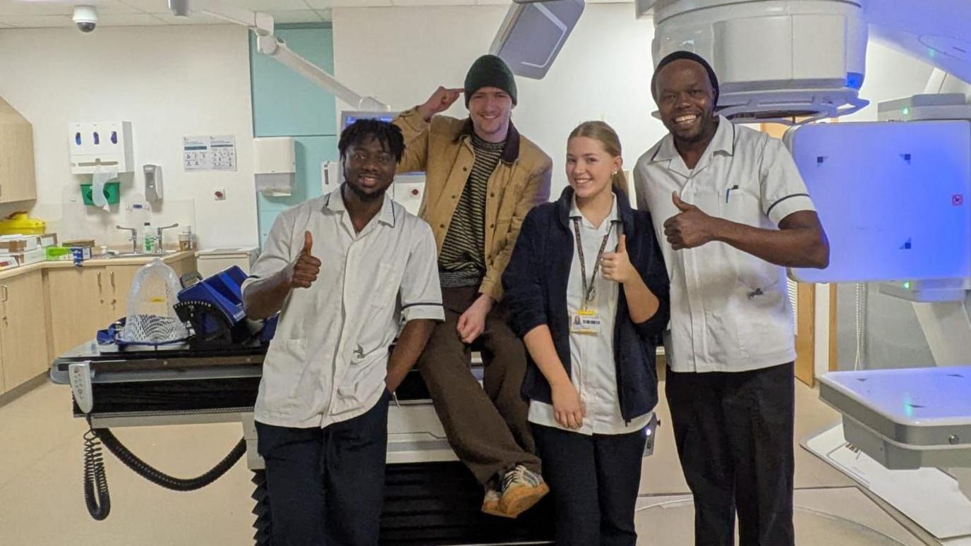 Arlo is in a radiotherapy room, sitting on a platform looking at the camera and smiling. He is pointing to his head and wears a dark green beanie hat, with a beige and black striped t shirt and camel coloured overshirt and brown wide legged trousers and rubber soled trainers. Standing either side of him are three medical staff, they all have their thumbs up and are smiling at the camera.