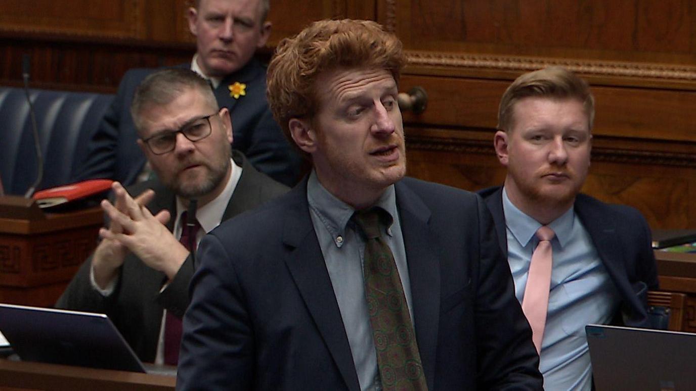 Matthew O'Toole speaks to the assembly in Stormont. He has ginger hair, wears clear glasses, and is wearing a dark coloured suit. Behind him are three other MLAS who are listening to him speak -Justin McNulty, Colin McGrath and Daniel McCrossan.