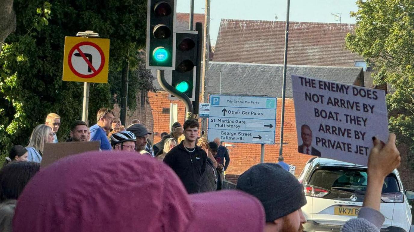 A counter-protester holds a sign that reads "the enemy does not arrive by boat, they arrive by private jet". The sign has a picture of Prime Minister Keir Starmer on it. Other protesters are gathered around the area.
