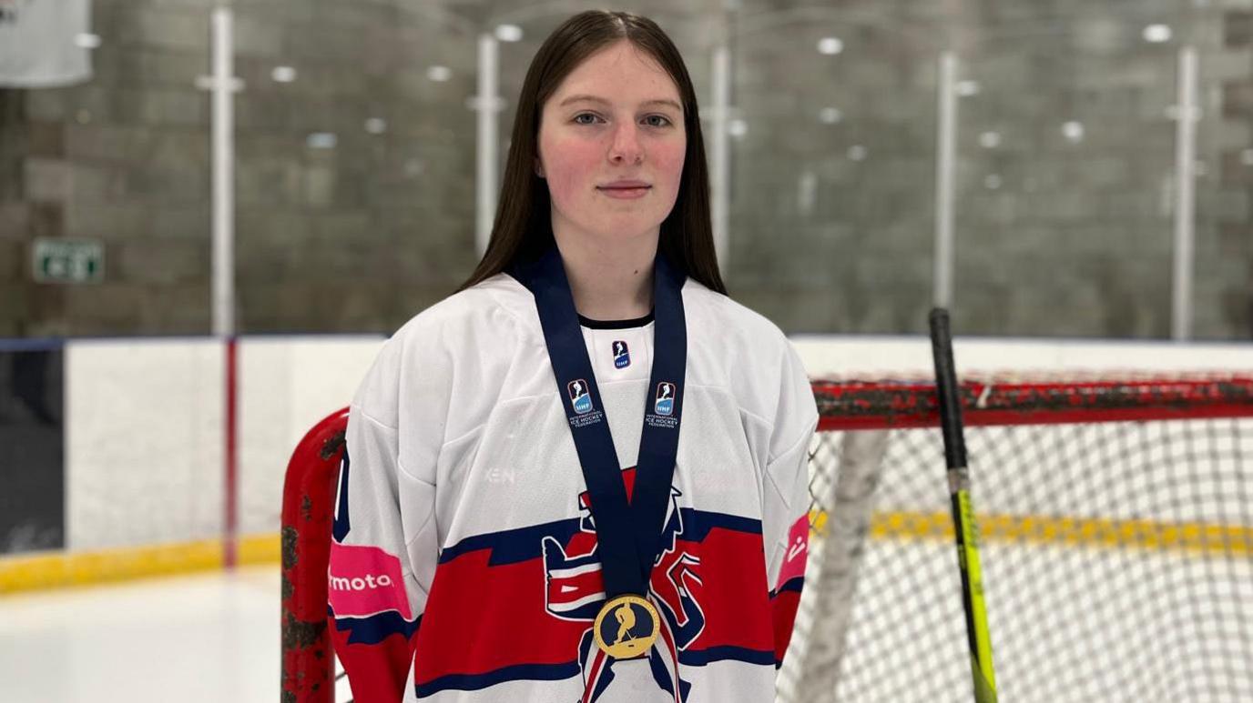 A young woman with dark hair in a Team GB ice hockey top with a medal around her neck stands in front of a hockey net