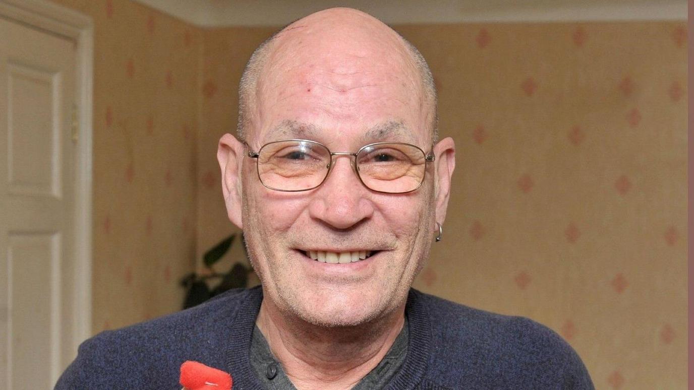 David Newton, wearing a grey sweater over a button-up T-shirt, and metal-framed spectacles, smiles as he holds up a red presentation box embossed with a gold National Blood Service logo and the figure "100" in one hand. In his left ear is an earring. In the other hand is a red soft toy mascot of a blood drop. He is inside what looks like a living room. 