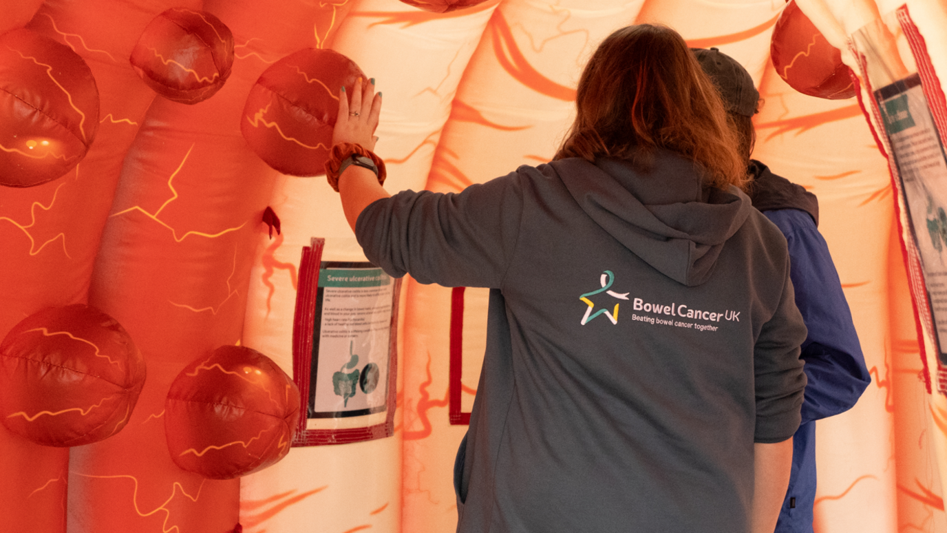 Two people with their backs to camera, one wearing a grey hoodie with the Bowel Cancer UK logo on the back, walking through a large, plastic pop-up tunnel simulating the inside of the bowel with large red round polyps