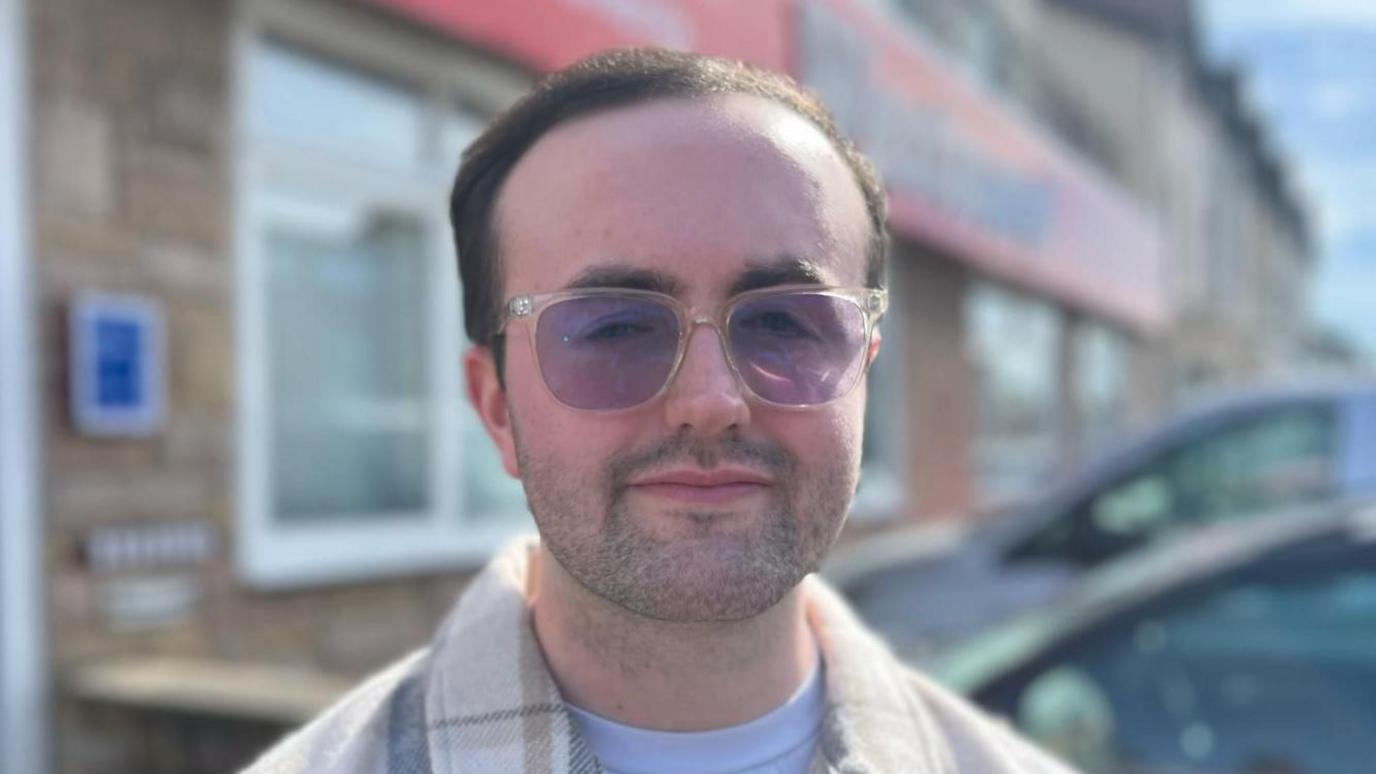 A close-up of Jordan Robbins, who has short hair and a short beard. He is wearing tinted glasses and a white t-shirt with a checked shirt over the top. The blurred background features a building and two parked cars.