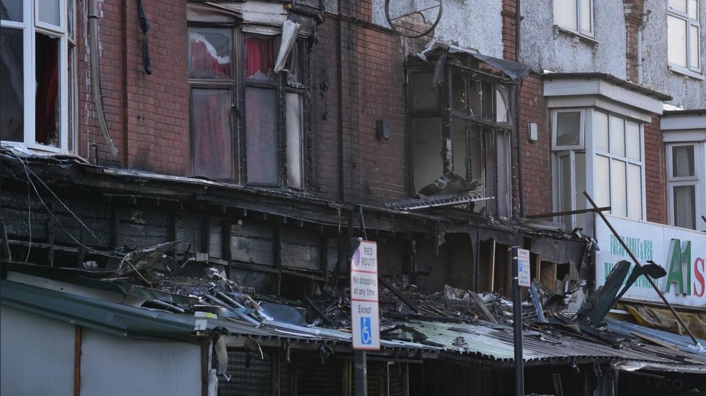 Flats above the shop have broken windows and blackened frames after the fire spread to homes above. The walls are also black and charred. The shop awnings are damaged and there is fire debris on them. Road signs are also visible from the pavement.