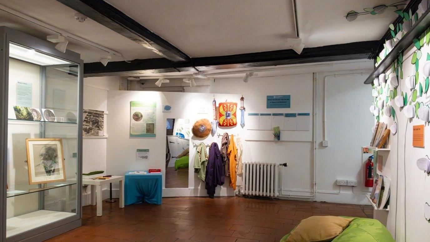 A white room with black faux beams on the ceiling. Left wall features a display case with exhibits and a small table for interactive work. The back wall has clothes to try on and a mirror. The right wall has a bookshelf and a display with writing on leaf-shaped paper. 