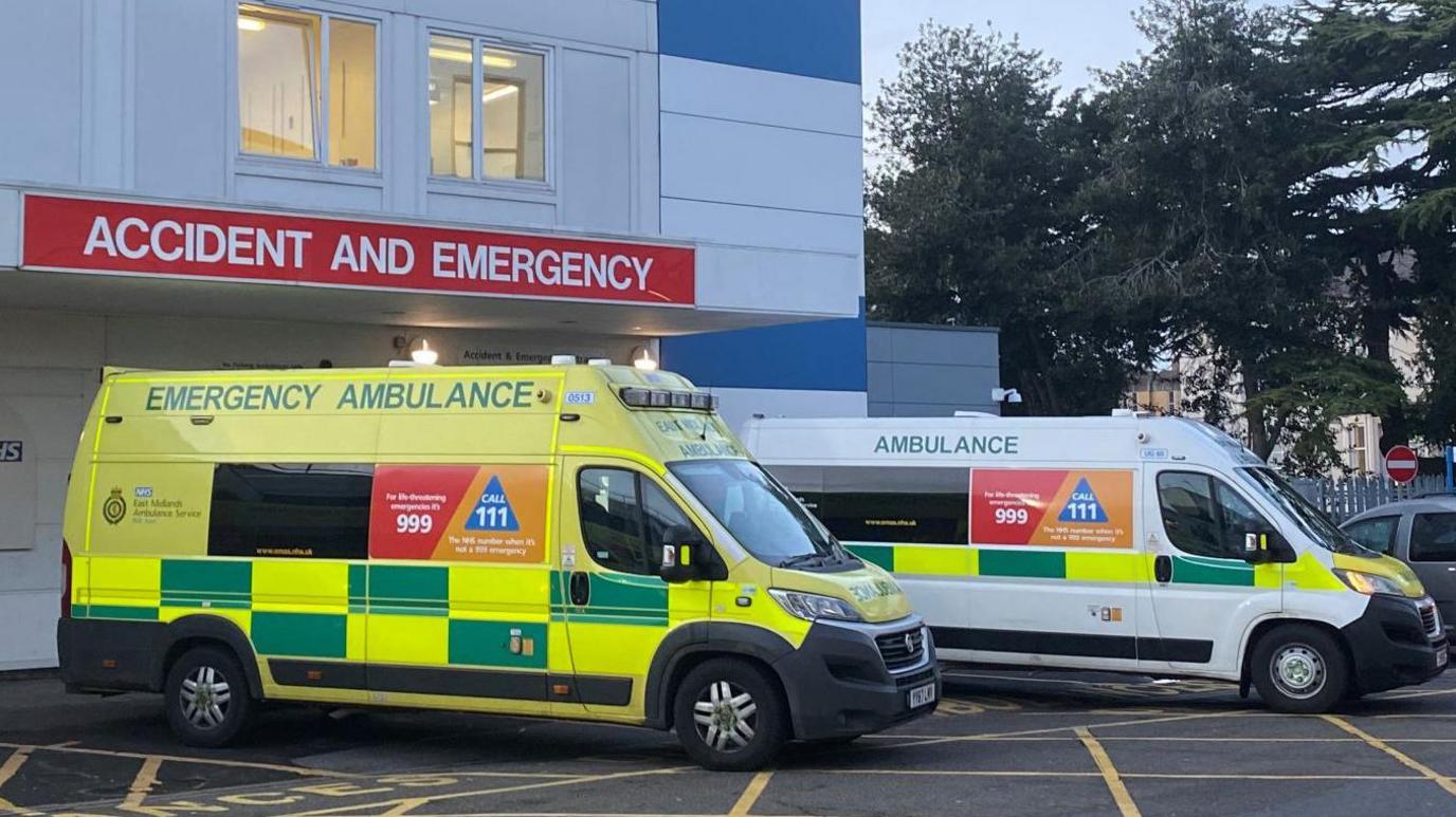 Two ambulances outside an emergency unit of a hospital. There is a sign on the mostly white building saying accident and emergency.