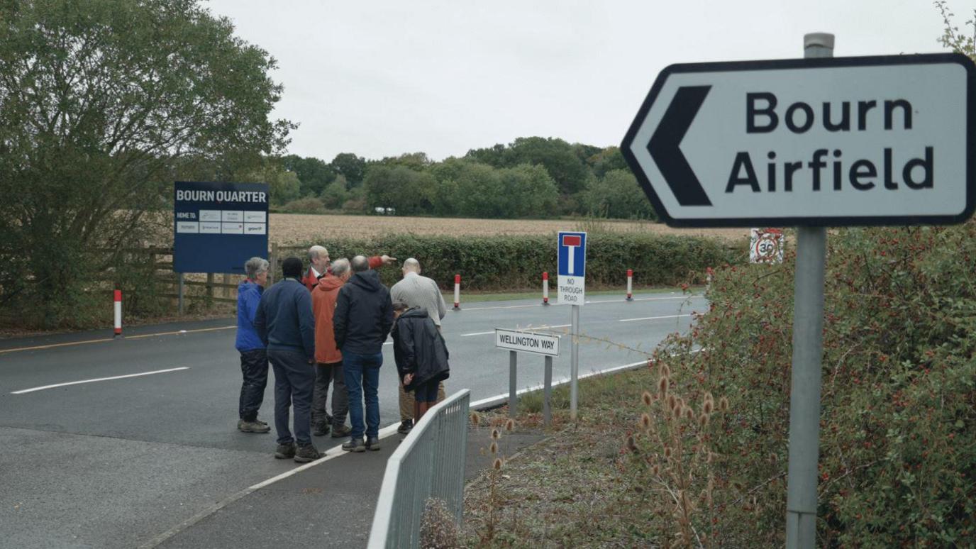 A group of people, including inspectors, look out over Bourn Airfield, with a sign in the foreground denoting the location.