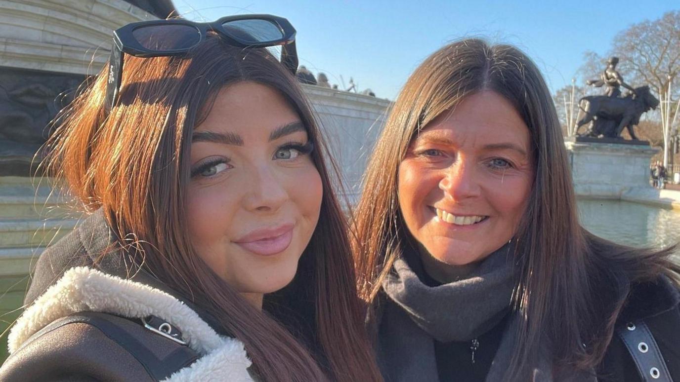 Debbie and Hollie Mytton stand smiling at the camera on a sunny day. Both have long brown hair parted in the middle