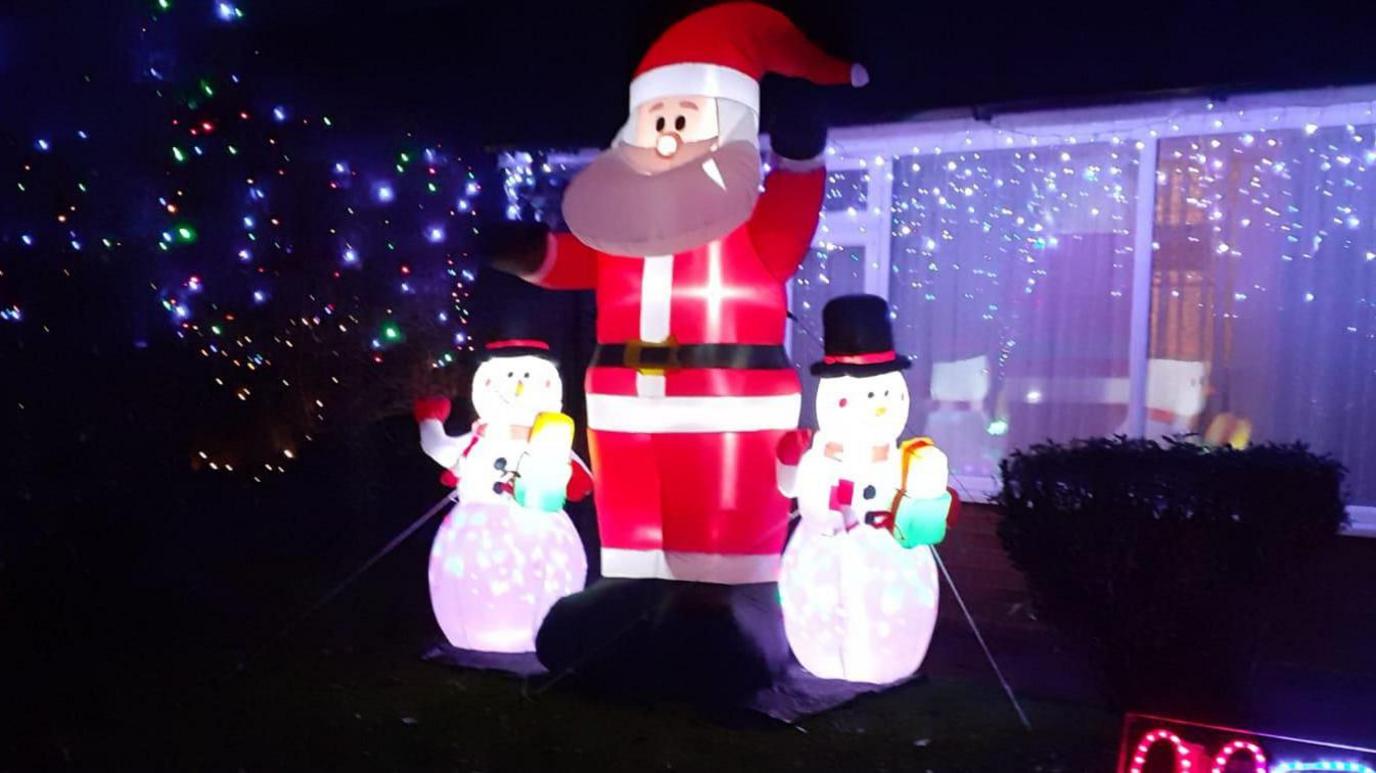 A large blow up Santa Claus decoration with two smaller snowmen next to it after pictured lit up in front of a home. More Christmas lights are decorated on the home along with a bush.