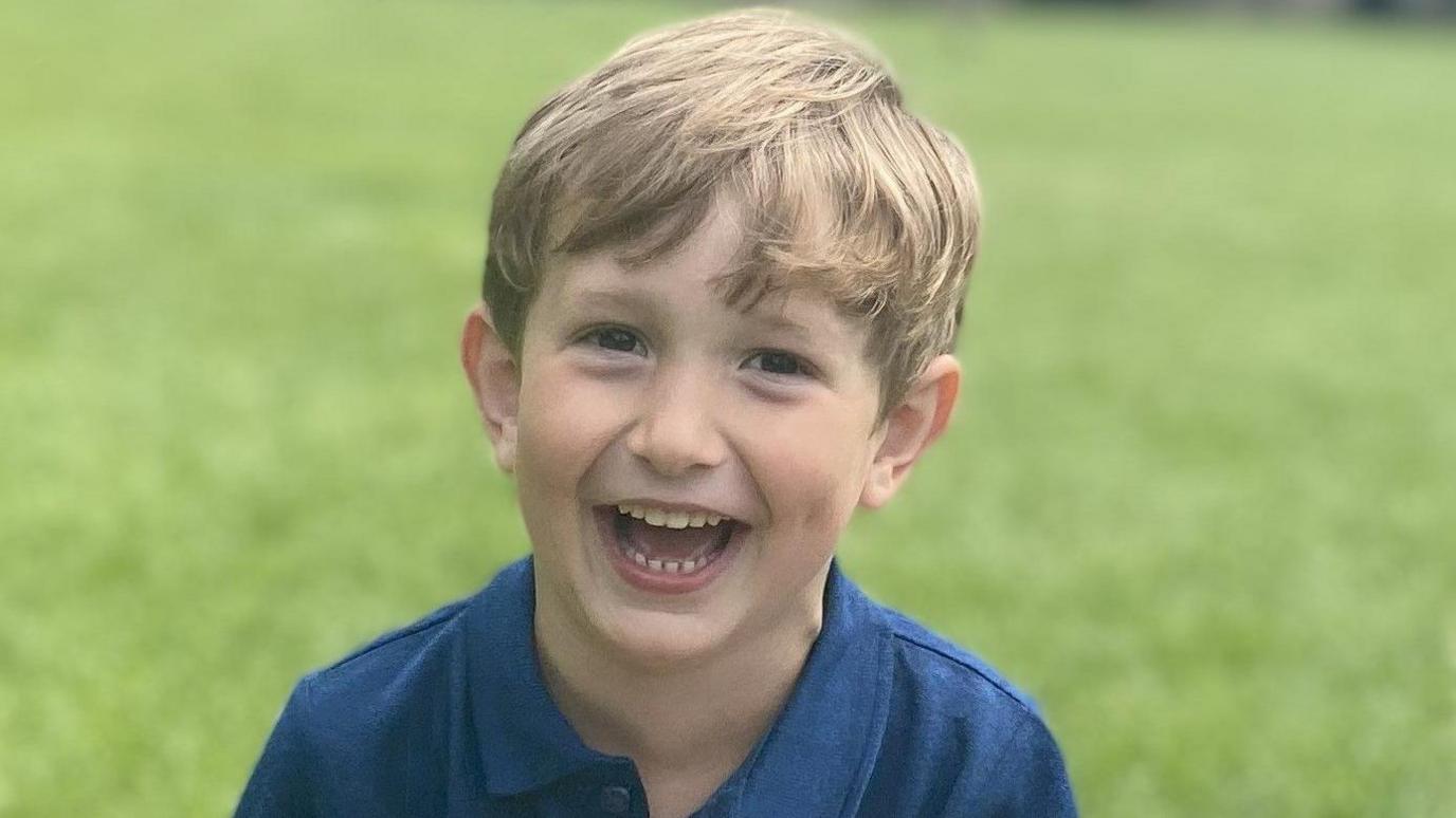 Benedict, a young boy with blond hair, is on a lawned space. He is wearing a blue polo shirt and is laughing into the camera.