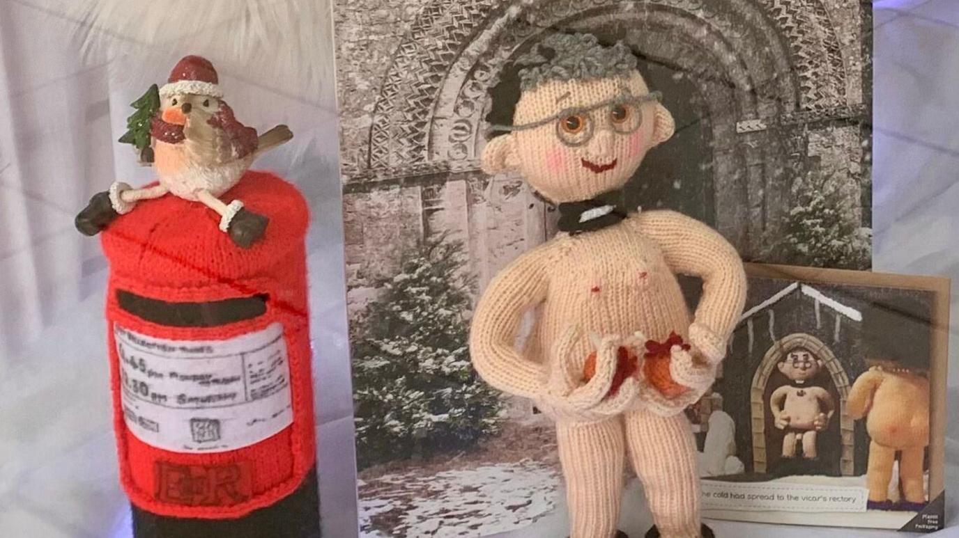 A nude knitted vicar stood by a photo of a church and a knitted post box with a robin on top.