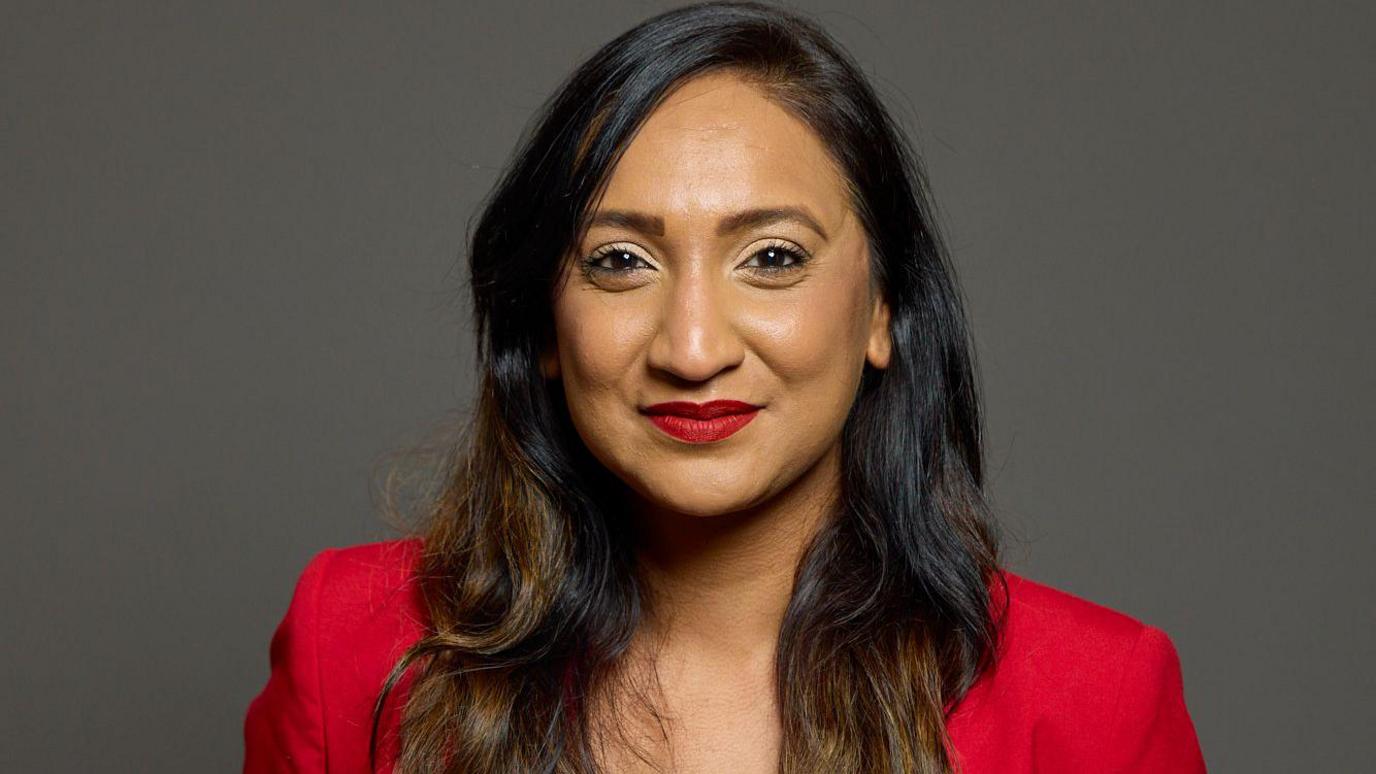 A head and shoulders image of a woman staring at the camera smiling. She has dark hair and wears a bright red jacket