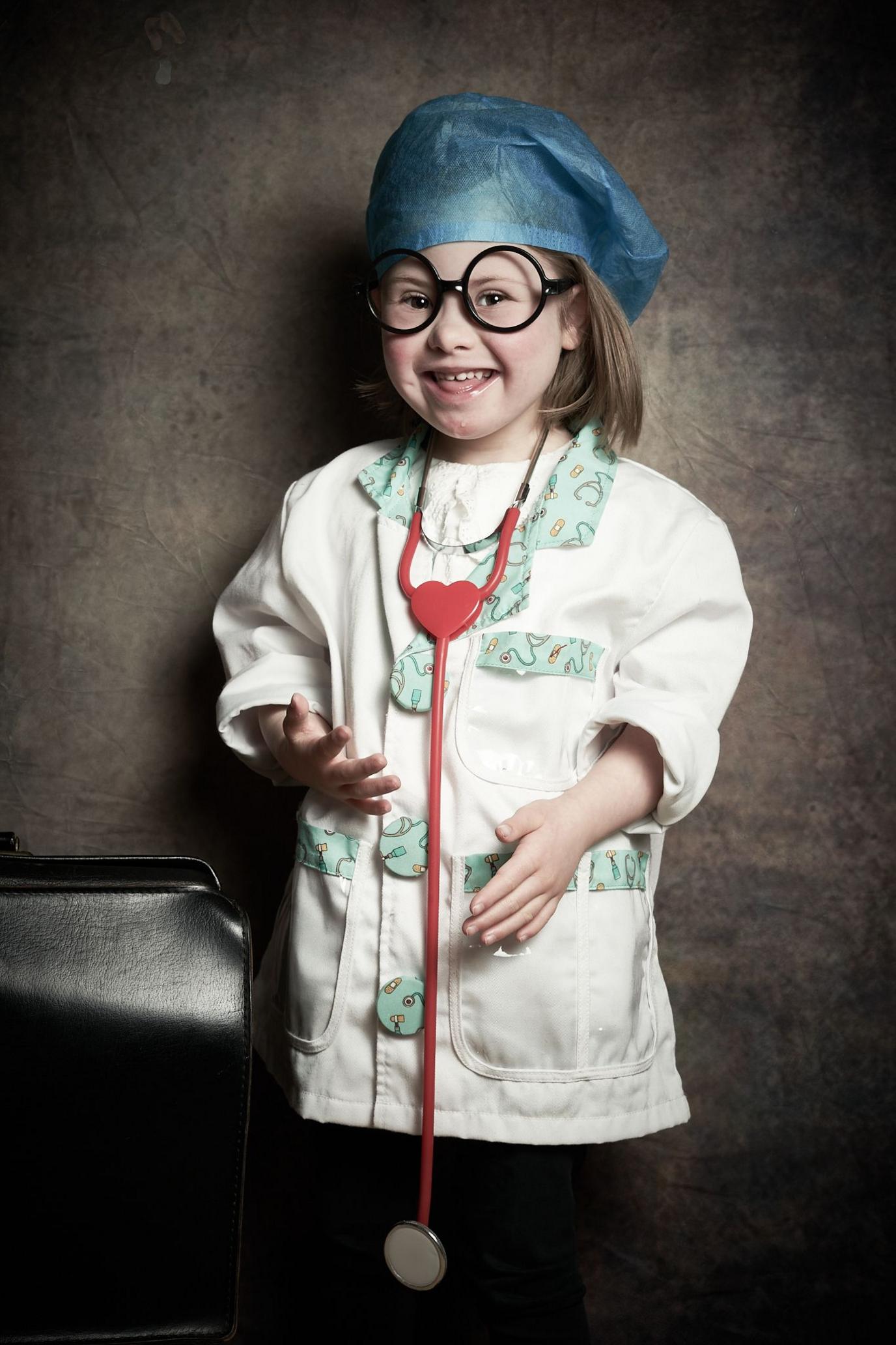 Layla, a young girl with shoulder length brown hair wearing a blue surgical hat, large round black-framed glasses, a child's nurse coat and large red stethoscope around her neck, smiles at the camera