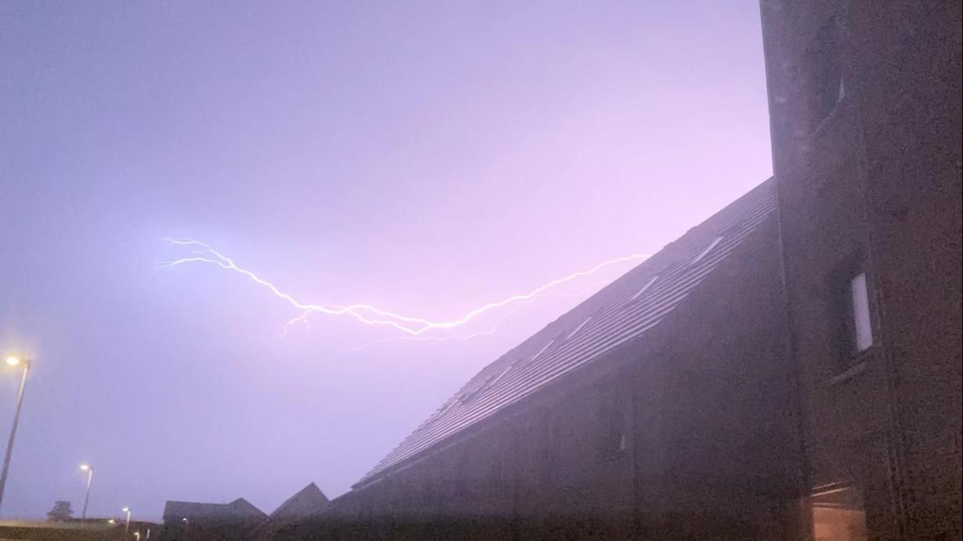 A purple sky with the silhouette of house roofs and a bright lightning strike in the sky.