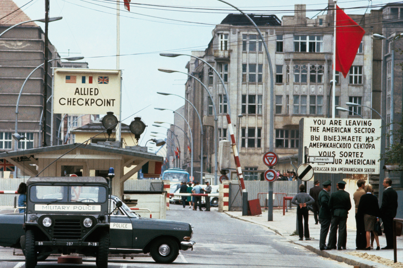 Archive photo of Checkpoint Charlie with military police vehicles parked in front of a wooden building with allied checkpoint written above it. Across the road several people mingle beneath a sign indicating that "you are leaving the American sector"