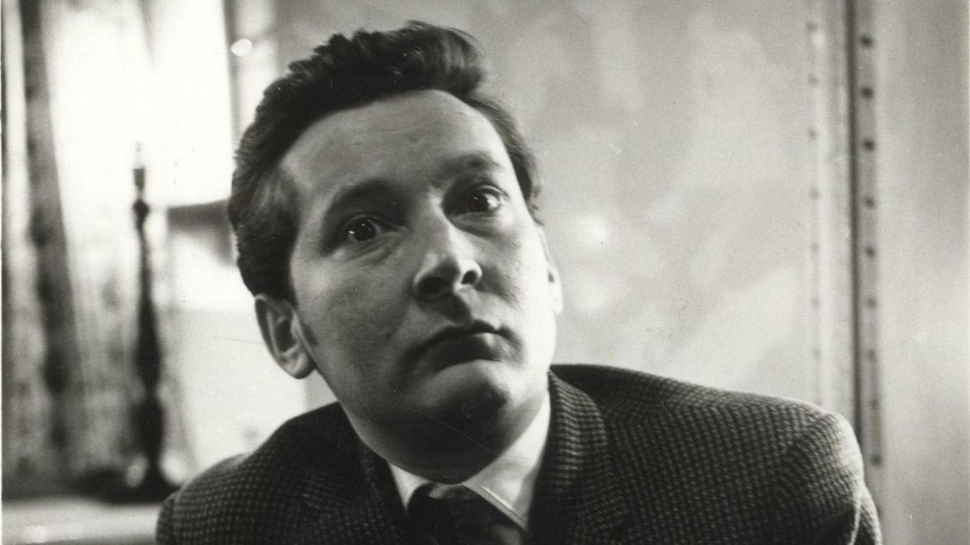 A black and white photo of Kenneth Williams shows him sitting in a room wearing a smart suit jacket, tie and white shirt