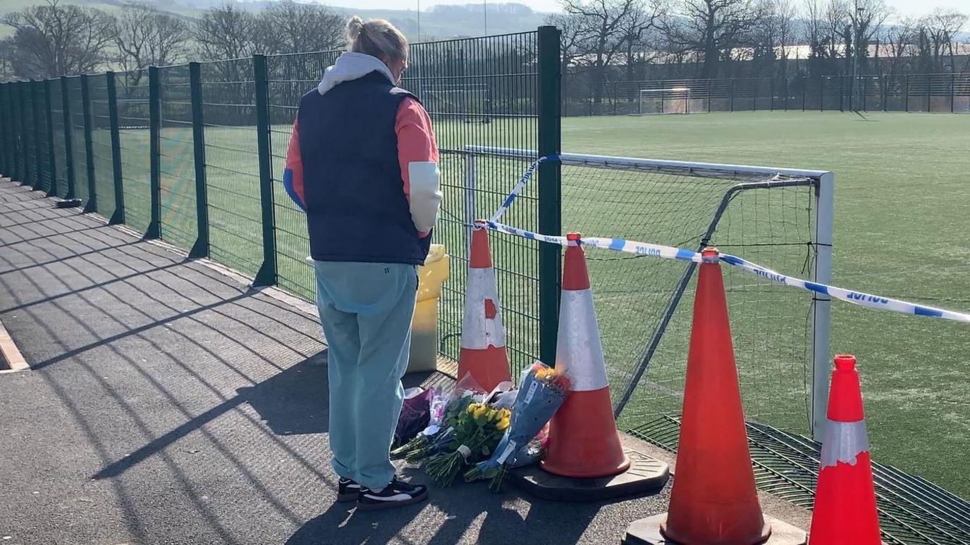 Six bouquets of flowers have been laid at the foot of a police cordon in front of the sports pitch. A woman wearing a bright hoodie and dark gilet stands with her back to the camera, bowing her head. A panel of green, metal fencing can be seen bent down on to the pitch, which has several five-a-side and full size football goalposts. The cordon is made up of orange traffic cones with blue and white police tape strung between them.