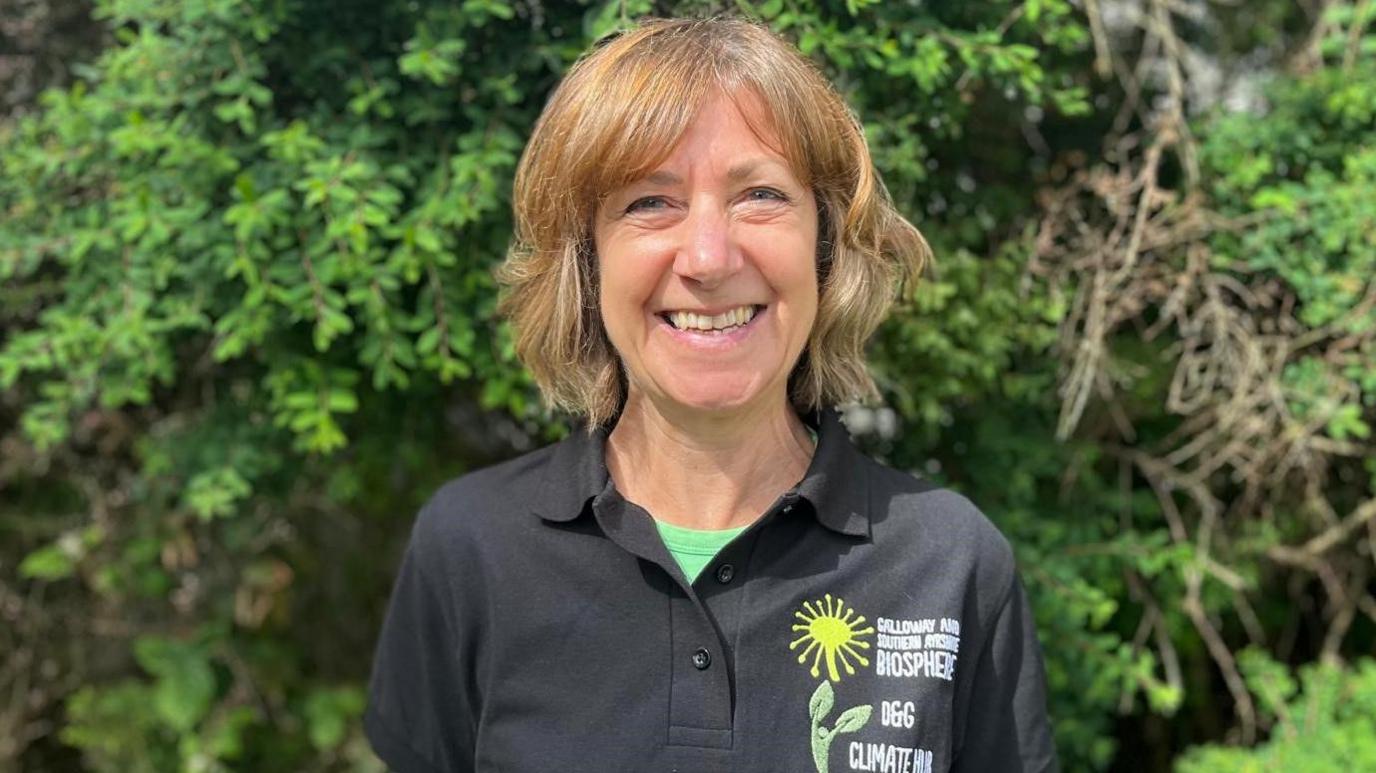 A smiling woman with short blonde wavy hair in a black button-necked T-shirt with a green T-shirt underneath