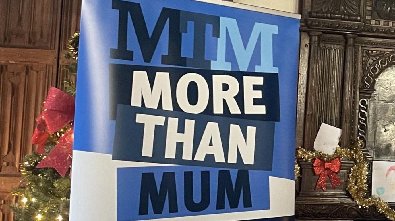 A sign that reads: "More Than Mum." The words are written on a blue and white background.