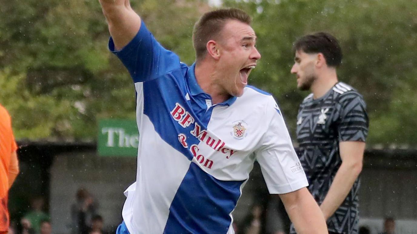 Harry Gibbs, in blue and white quartered shirt, wheels away with an arm up to celebrate a goal, while an opponent in a black and grey shirt looks disappointed behind him