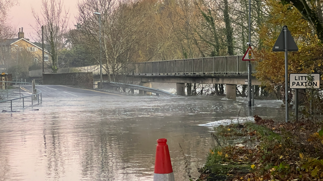 Flood water has taken over a road which can no longer be seen. Towards the back of the image the road reappears as it goes over a bridge. To the right of the image is a sign for Little Paxton. The sunrise is pink in the sky.