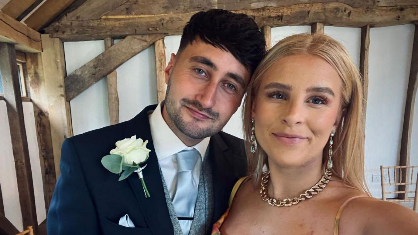 Oliver Dent and his partner Caitlin Phillips-Diggons stood in an old property which has white walls and wooden studs. Oliver is wearing a navy suit with a white flower pinned on the front, while Caitlin is wearing a dress with a gold chain around her neck. She has shoulder length blonde hair and both are smiling.