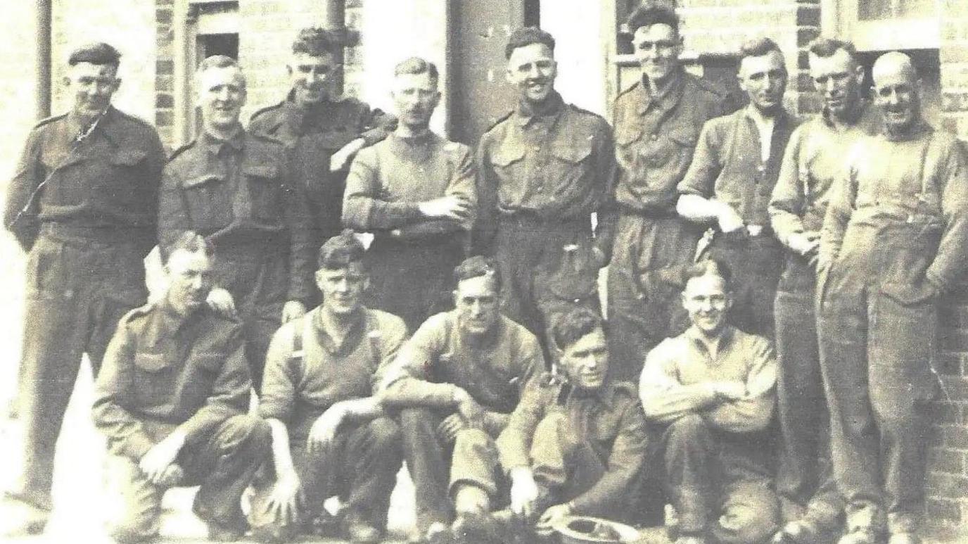A black and white photo of fourteen men standing outside a brick building, nine are standing in a row behind five others who are squatting, sitting or kneeling down. They're wearing army uniform and looking towards the camera.