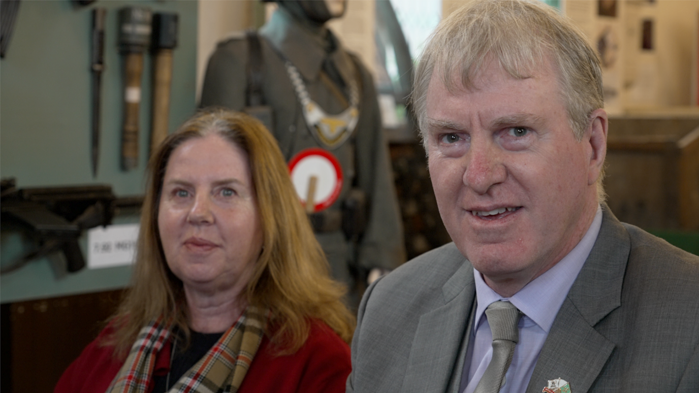 Mr Le Druillenec's grandchildren Peter and Linda smile at the camera. He is wearing a grey suit jacket and tie and she is wearing a red coat and checked scarf. Behind them is a World War Two display featuring a military uniform and a gun.