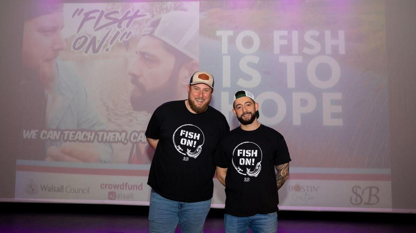 Two men stand in front of a large projector screen. The men are wearing black t-shirts with the words 'Fish On!' written on the front.