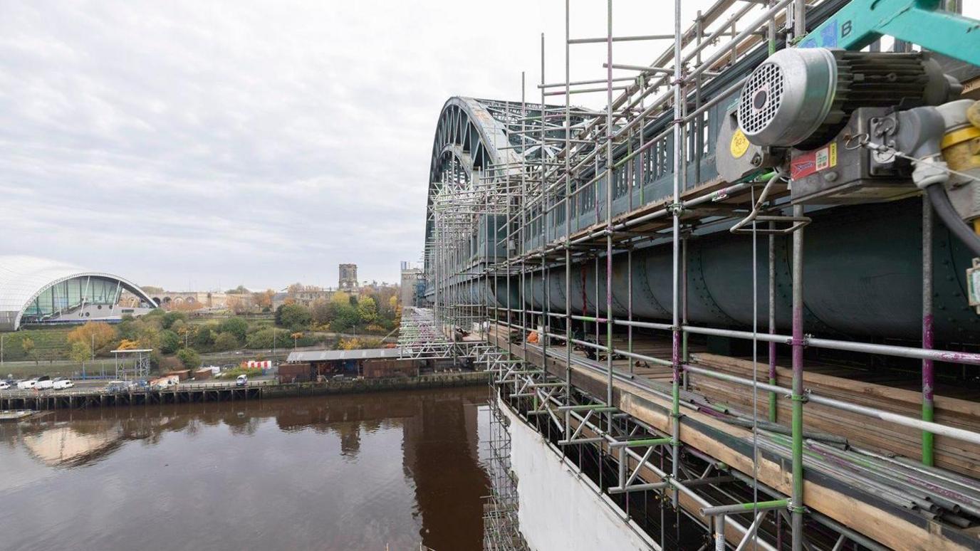 Scaffolding at the bottom of the arched Tyne Bridge. Gateshead's Glasshouse can be seen across the Tyne river, surrounded by colourful autumn trees. 