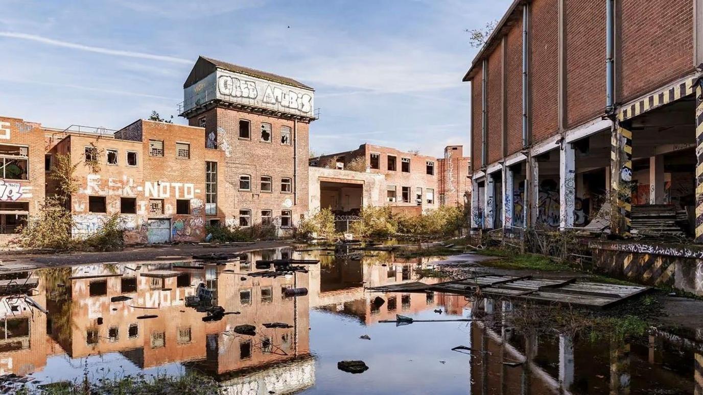 Old industrial brick buildings. The land between two separate blocks is muddy and water covers the floor, reflecting the buildings in it.