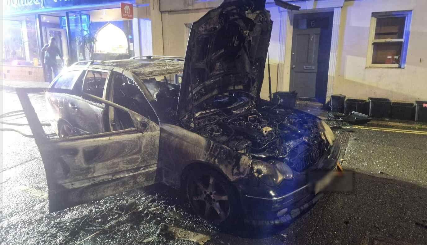 A car - possibly a Skoda - with its bonnet and a car door open and the charred remains of the engine and the interior of the car along with flame marks on the exterior suggesting that the car has been on fire, with a house behind along with a line of what appear to be household waste vestibules which appear to of the food caddy genre.