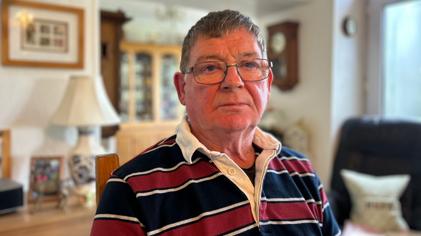 Alun Owens yn gwisgo sbectol a chrys streipiog coch a du gyda choler gwyn.