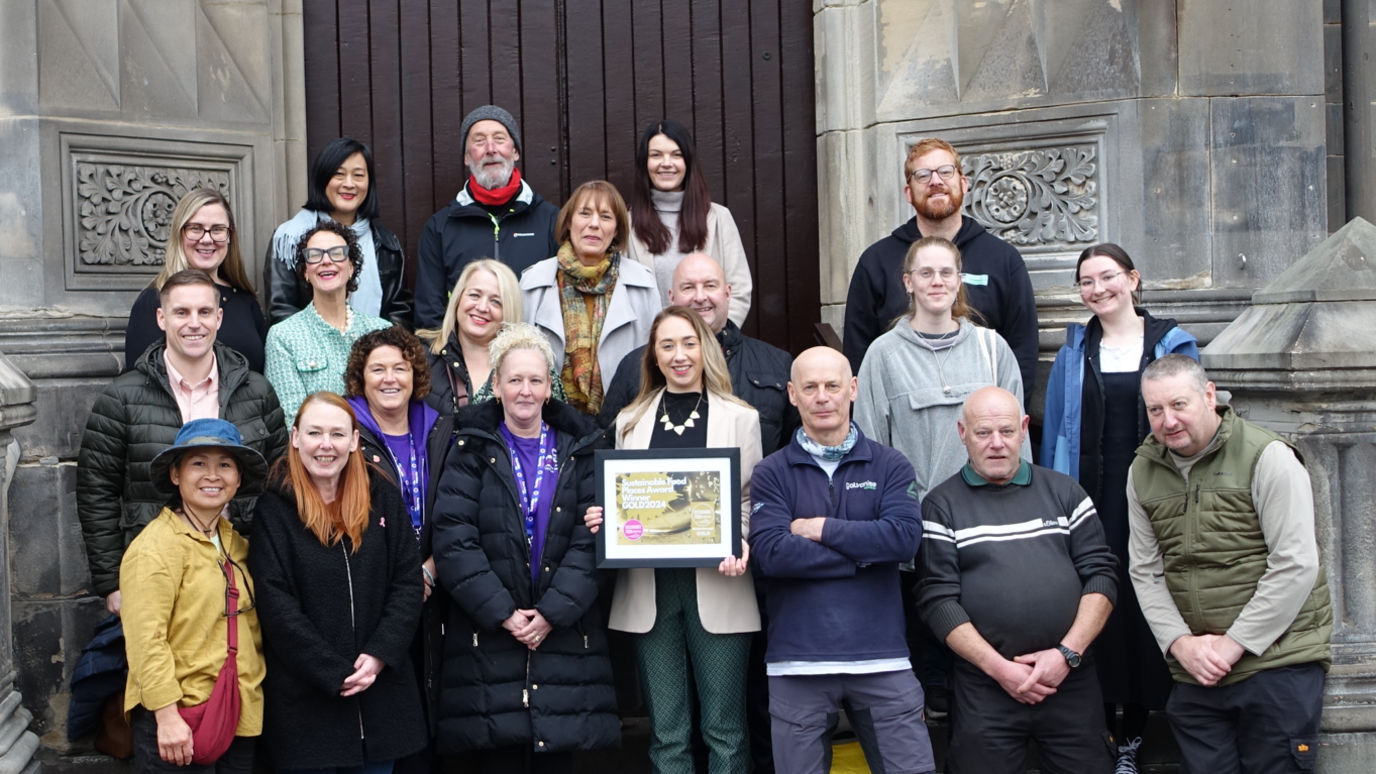 Dozen of people pose in front of a building's entrance. One person in the middle is holding a framed certificate. They are all smiling at the camera.