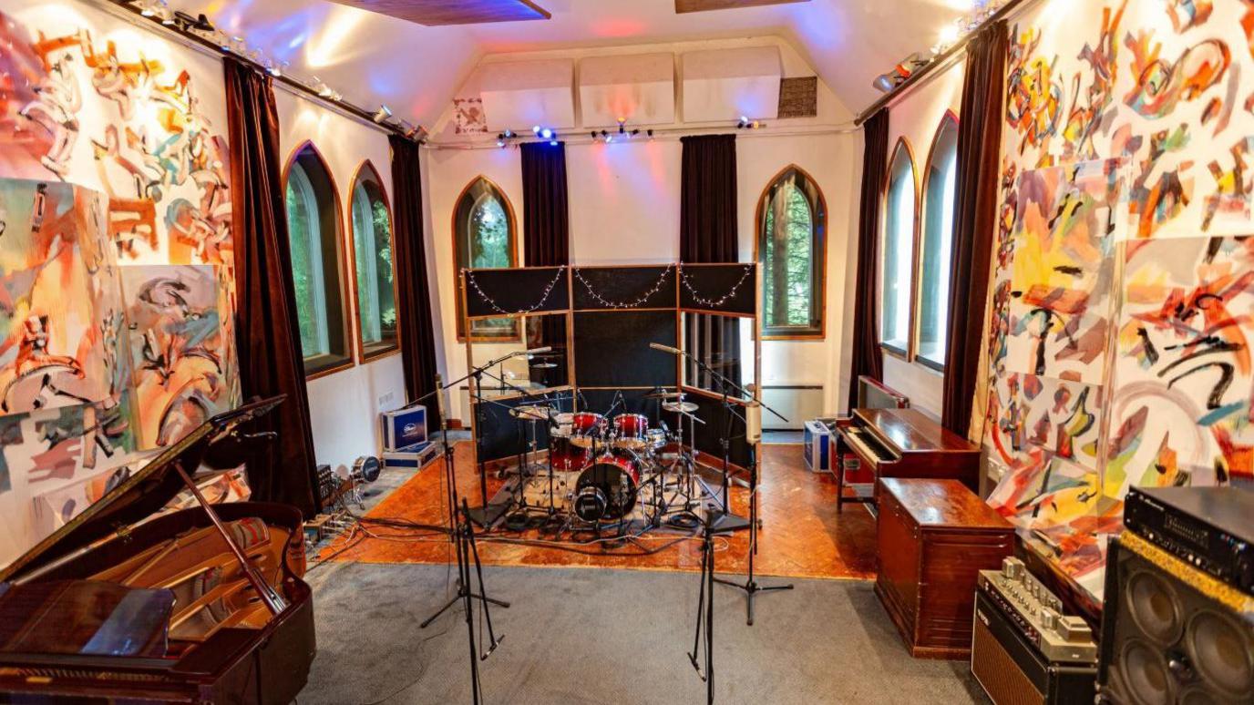 A recording studio inside a room with arched windows. In the centre is a red drum set surrounded by microphones and sound panels. A brown grand piano is on the left and another smaller piano is on the right. Abstract paintings hang on the walls and lights are strung across the ceiling.