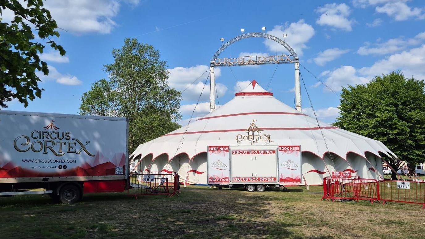 A circus big top, white with red trim. A nearby lorry, also white with some red patterning on the side, is emblazoned with a logo reading "CIRCUS CORTEX".