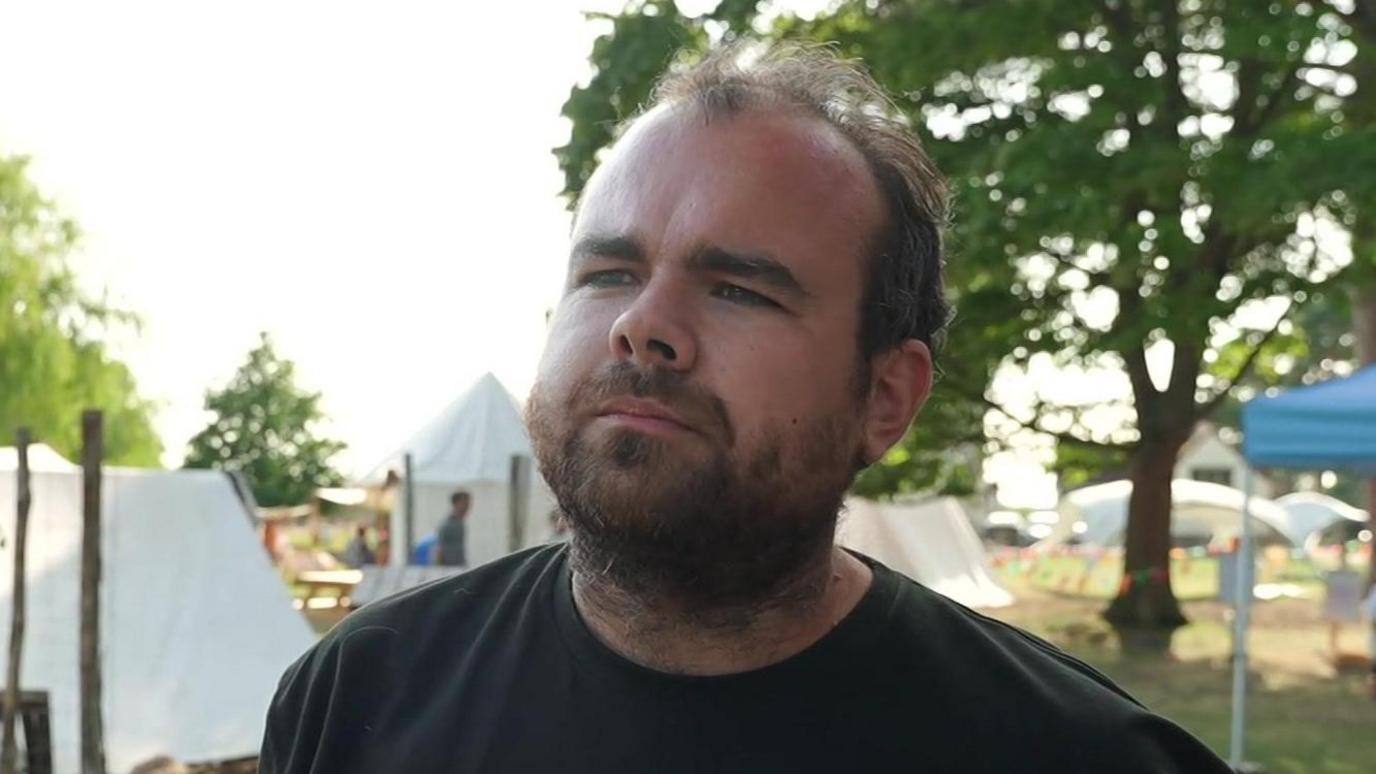 Josh Ward looks to the left of the camera and he has a concentrating expression on his face. He has thinning dark hair and a thick beard. He wears a black T-shirt.