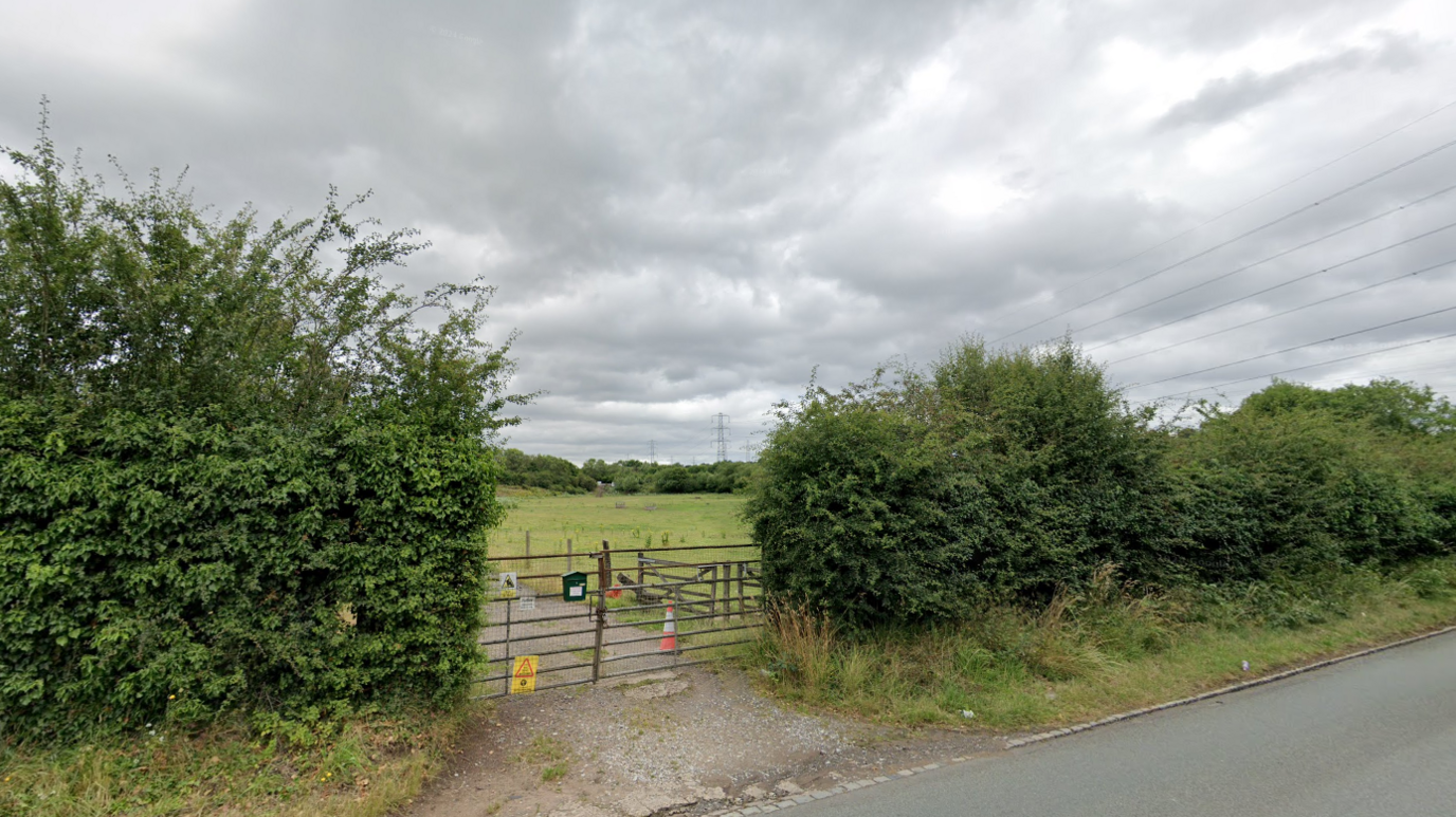 A gate leads off a road into a field. Hedges separate the road from the field. Trees can be seen in the distance at the boundary of the field.