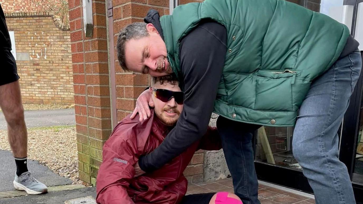 A man sitting outside on the ground hugging a man who is standing up and bent over him. The man on the ground is wearing a red waterproof zip-up jacket and colourful sunglasses. He has bright pink tape on his knee. The man embracing him is wearing blue jeans, a navy hoodie and a green bodywarmer. He has dark, greying hair.