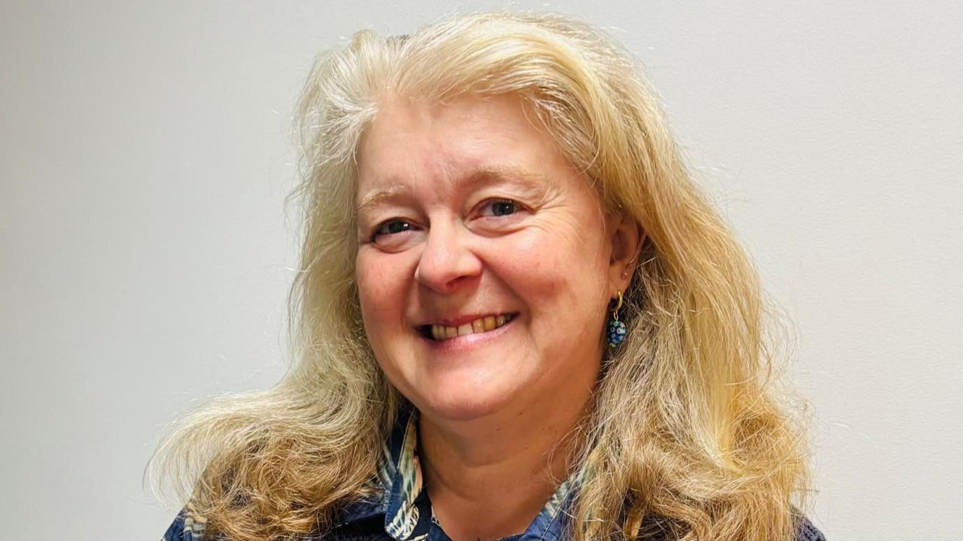 Sarah Daniels looks towards the camera. She is smiling and has long blonde hair. She is wearing a blue patterned shirt with a blue jumper over the top.