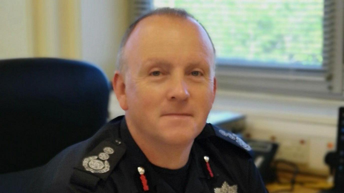 Mark Preece wearing his uniform in a corporate environment, smiling slightly at the camera. He is a middle aged man and is balding. There is a window looking out onto greenery behind him.