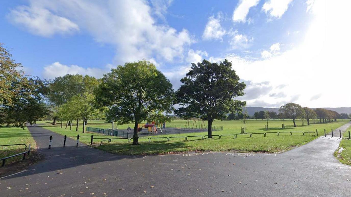 A wide view of a green space in Eston Recreation Ground. Grassy areas are intersected with grey pavement. A small children's play park is seen in behind two trees.