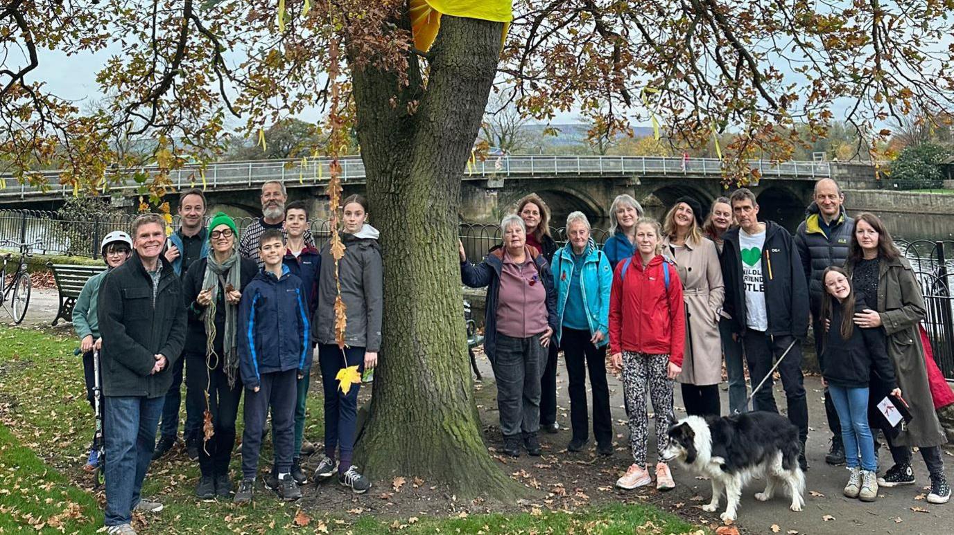 Campaigners against the felling of an oak tree in Otley stand by it, there are about 20 people and a dog, and a bridge can be seen in the background