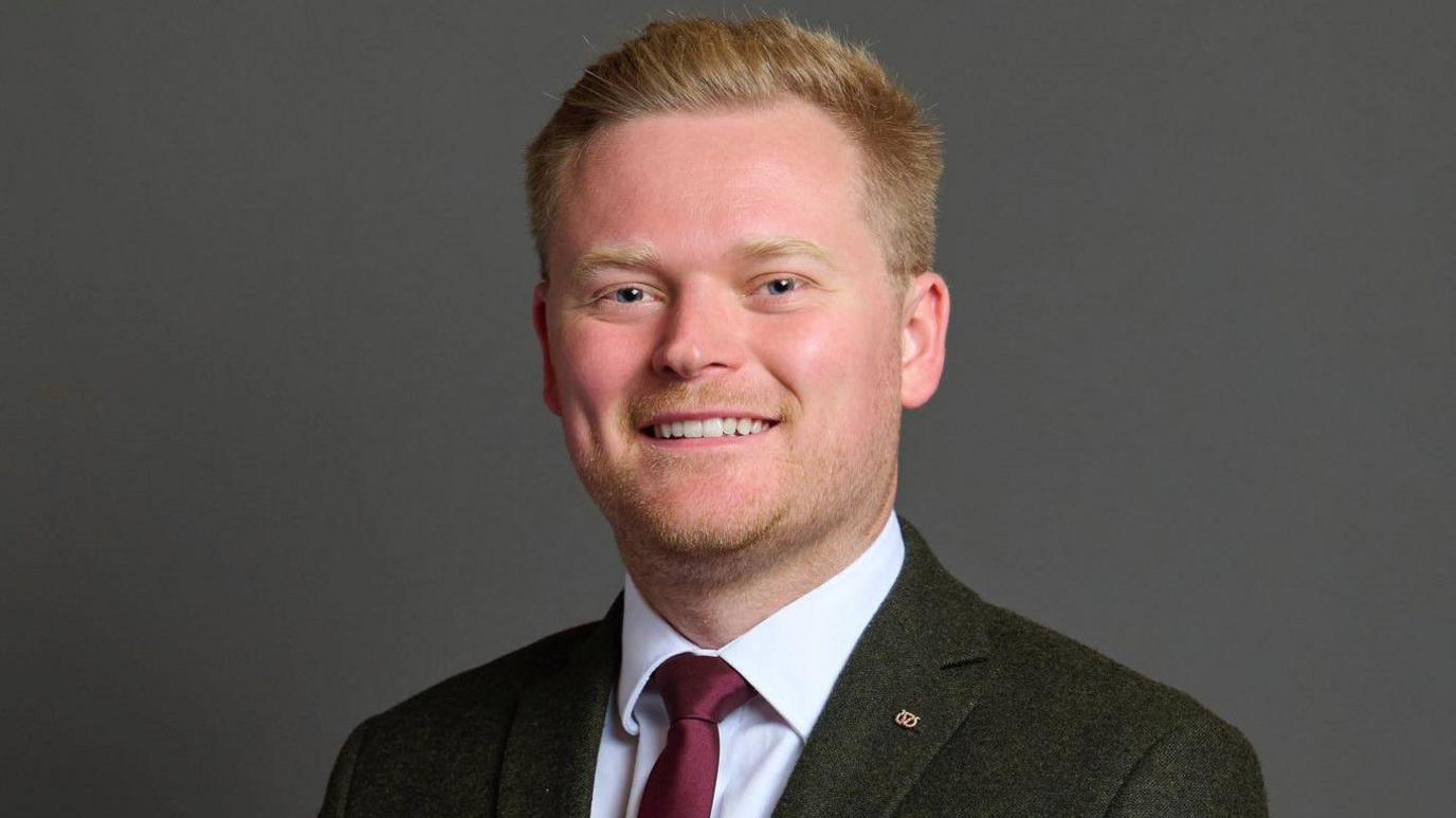 Photograph of Joshua Newbury MP, wearing a grey suit and maroon tie. 