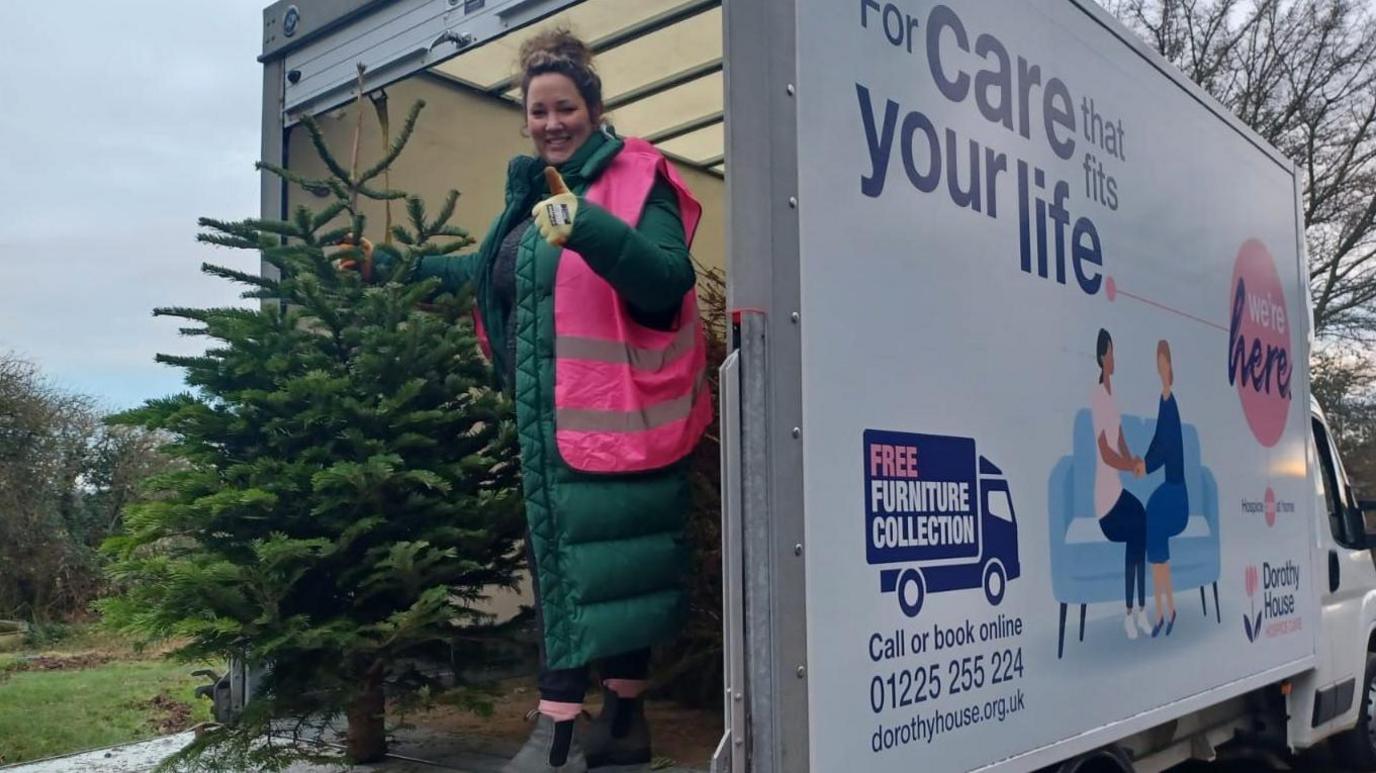 A woman standing in the back of a van with a used Christmas tree. The van has a Dorothy House graphic on the side. The woman is wearing a long green puffer coat and a high vis pink vest. She has dark hair tied up in a bun. She is looking at the camera and smiling while holding her thumb up. 