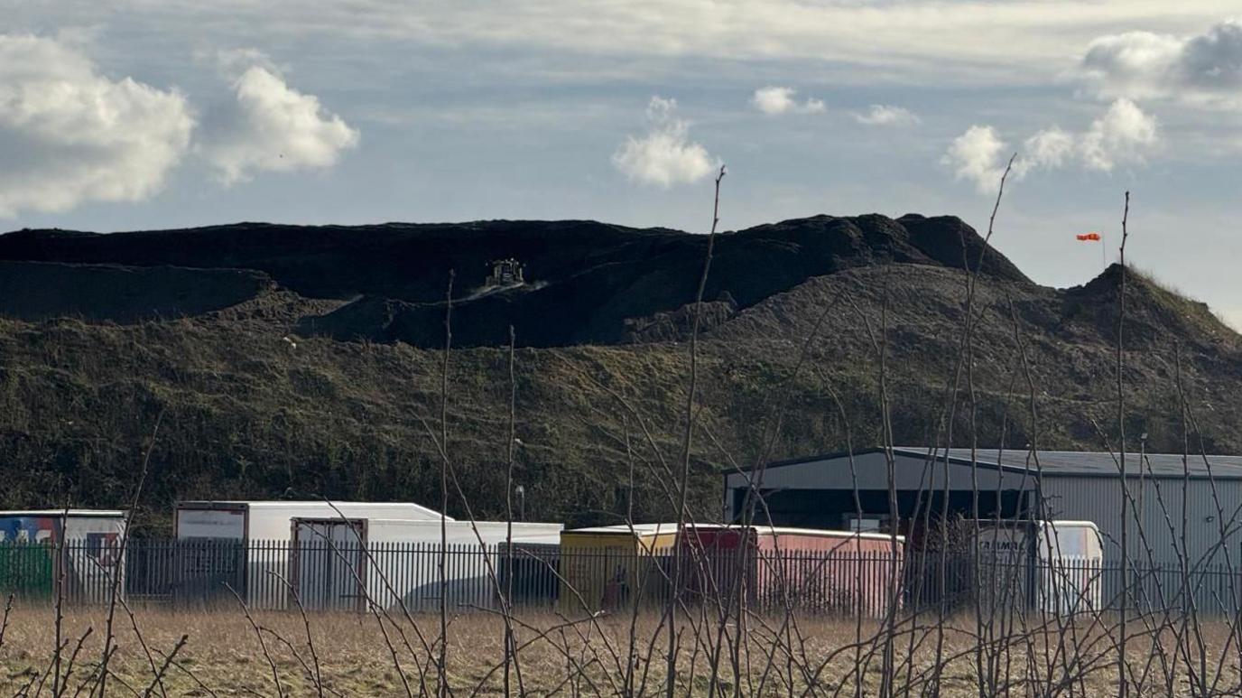 waste mound on Simonswood Industrial site looms over sheds and lorries nearby